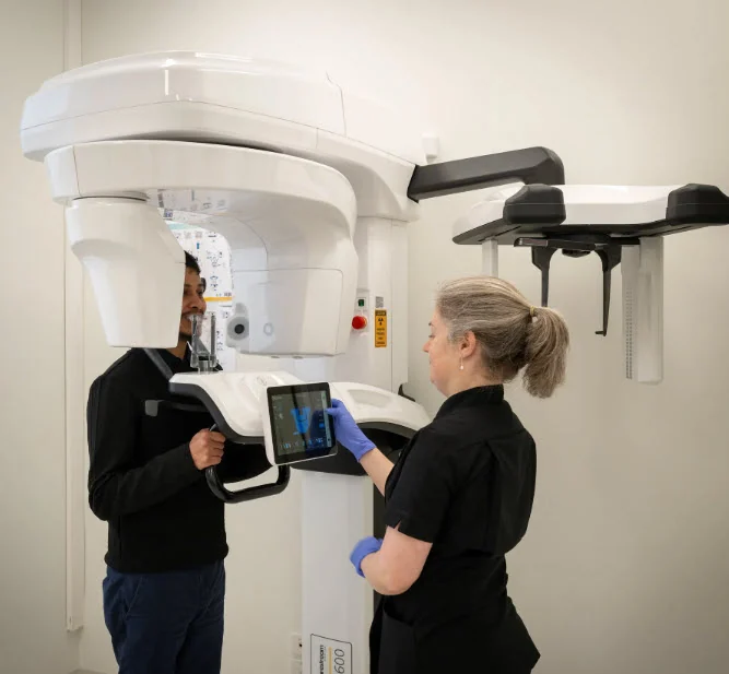 A patient positioned in the 3D cone beam scanner while an assistant sets up the scan at the console